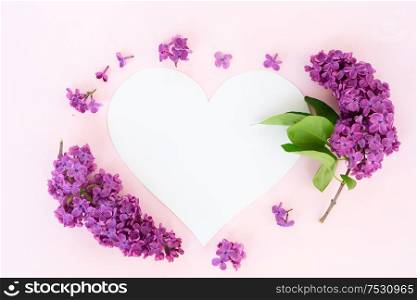 Fresh lilac flowers over pink background with copy space on heart paper note, flat lay floral composition. Fresh lilac flowers