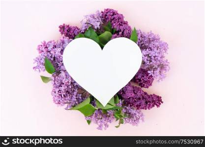 Fresh lilac flowers over pink background with copy space on heart shaped paper note, flat lay floral composition. Fresh lilac flowers