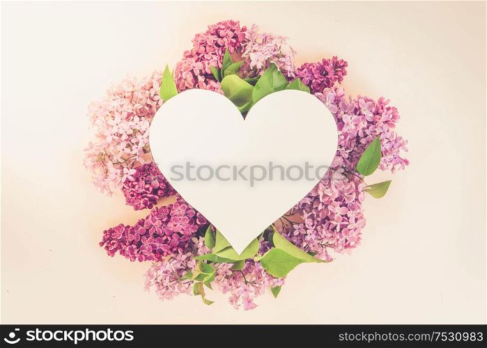 Fresh lilac flowers over pink background with copy space on heart shaped paper note, flat lay floral composition, toned. Fresh lilac flowers