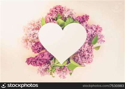 Fresh lilac flowers over pink background with copy space on heart shaped paper note, flat lay floral composition, toned. Fresh lilac flowers