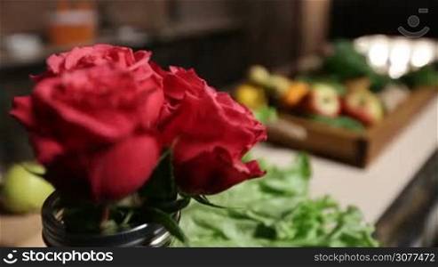 Fresh market fruits and vegetables in wooden tray lying on the table in household kitchen. Foreground bouquet of red roses in water-glass. Selective focus. Healthy eating and dieting concept.