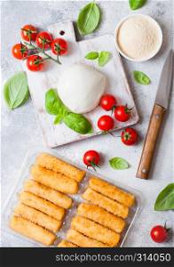 Fresh Mozzarella cheese on vintage chopping board with tomatoes and basil leaf and tray with cheese sticks on stone kitchen background.