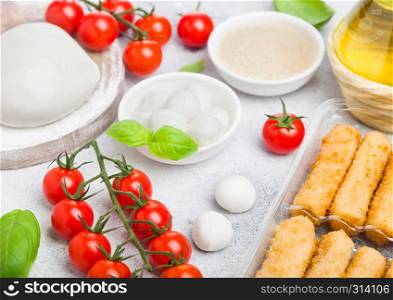 Fresh Mozzarella cheese on vintage chopping board with tomatoes and basil leaf with olive oil and tray with cheese sticks on stone kitchen background.
