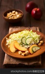 Fresh nectarine, cucumber and iceberg lettuce salad on wooden plate with homemade croutons on the side, photographed on dark wood with natural light (Selective Focus, Focus on the cucumber slices in the middle of the plate)