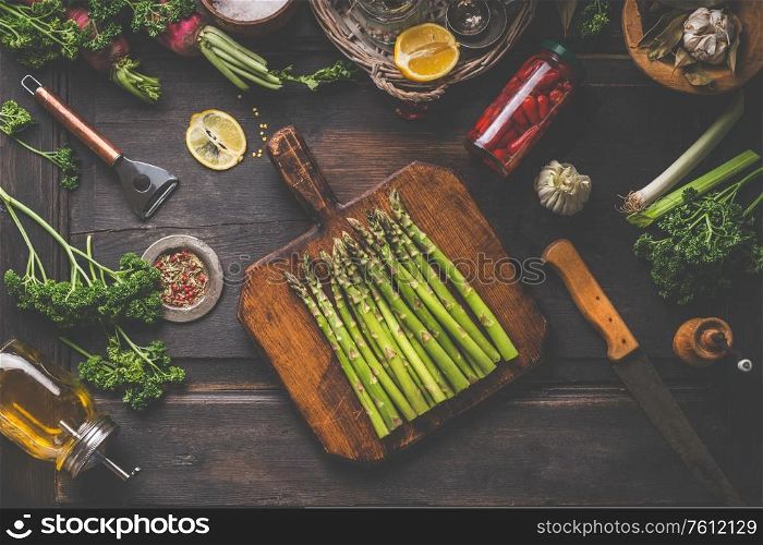Fresh organic asparagus on wooden cutting board. Copped herbs, olive oil, lemon and seasonings. Preserved hot peppers in glass jar on dark rustic background, top view. Healthy lifestyle. Home cooking