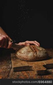 fresh organic bread on a wooden board cut a man&rsquo;s hands on a dark background and against the background of an old wooden table. fresh organic bread cut a man&rsquo;s hands