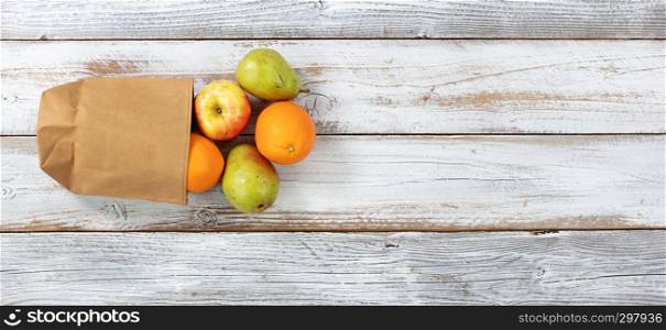 Fresh organic fruit pouring out of recyclable paper bag on white rustic wooden background