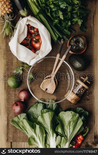 Fresh organic ingredients for salad making and wooden spoons with glass salad bowl on rustic background, top view. Flat lay with place for text. Vegan and healthy food concept