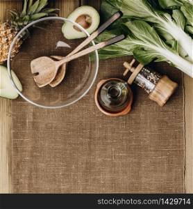 Fresh organic ingredients for salad making and wooden spoons with glass salad bowl on rustic background, top view. Flat lay with place for text. Vegan and healthy food layout