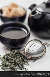 Fresh organic loose green tea with strainer infuser and black teapot and cane sugar on white table background.