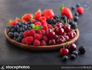 Fresh organic summer berries mix in round wooden tray on black kitchen table background. Raspberries, strawberries, blueberries, blackberries and cherries. Top view