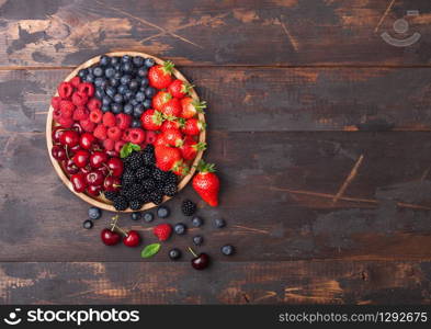 Fresh organic summer berries mix in round wooden tray on dark wooden table background. Raspberries, strawberries, blueberries, blackberries and cherries. Top view