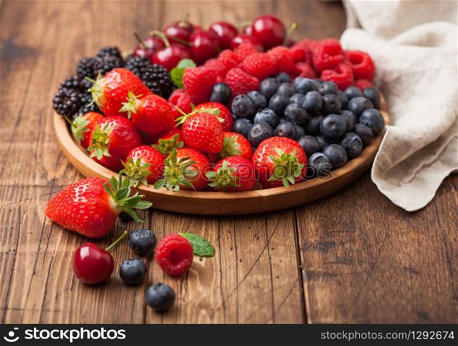 Fresh organic summer berries mix in round wooden tray on light wooden table background. Raspberries, strawberries, blueberries, blackberries and cherries with linen kitchen towel. Top view