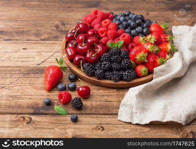 Fresh organic summer berries mix in round wooden tray on light wooden table background. Raspberries, strawberries, blueberries, blackberries and cherries with linen kitchen towel.  Top view