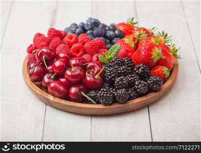 Fresh organic summer berries mix in round wooden tray on light wooden table background. Raspberries, strawberries, blueberries, blackberries and cherries. Top view