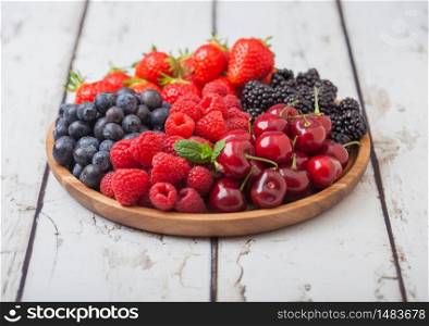Fresh organic summer berries mix in round wooden tray on white wooden table background. Raspberries, strawberries, blueberries, blackberries and cherries. Top view