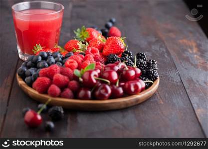 Fresh organic summer berries mix in round wooden tray with glass of juice on dark wooden table background. Raspberries, strawberries, blueberries, blackberries and cherries. Top view