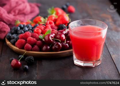 Fresh organic summer berries mix in round wooden tray with glass of juice on dark wooden table background. Raspberries, strawberries, blueberries, blackberries and cherries.  Top view