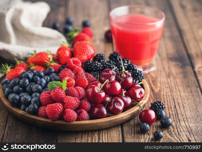 Fresh organic summer berries mix in round wooden tray with glass of juice on light wooden table background. Raspberries, strawberries, blueberries, blackberries and cherries. Top view