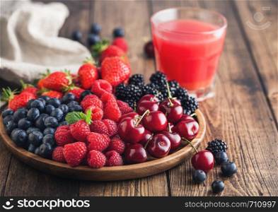 Fresh organic summer berries mix in round wooden tray with glass of juice on light wooden table background. Raspberries, strawberries, blueberries, blackberries and cherries. Top view