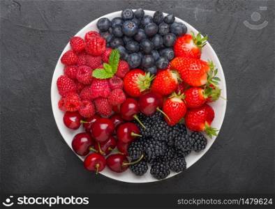 Fresh organic summer berries mix in white plate on black kitchen table background. Raspberries, strawberries, blueberries, blackberries and cherries. Top view