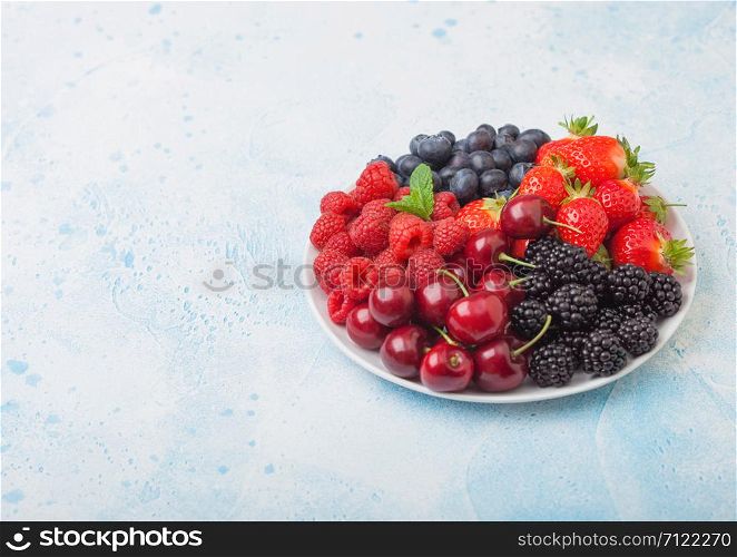 Fresh organic summer berries mix in white plate on blue kitchen table background. Raspberries, strawberries, blueberries, blackberries and cherries. Top view