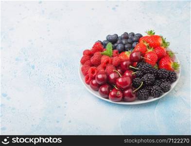 Fresh organic summer berries mix in white plate on blue kitchen table background. Raspberries, strawberries, blueberries, blackberries and cherries. Top view