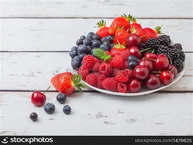 Fresh organic summer berries mix in white plate on light wooden table background. Raspberries, strawberries, blueberries, blackberries and cherries. Top view