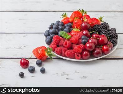 Fresh organic summer berries mix in white plate on light wooden table background. Raspberries, strawberries, blueberries, blackberries and cherries. Top view