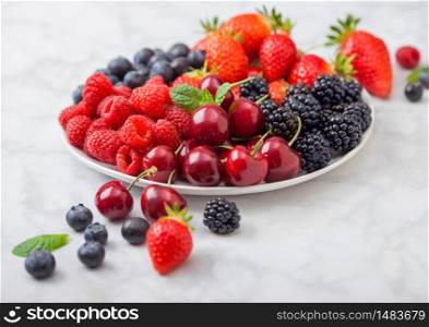 Fresh organic summer berries mix in white plate on marble background. Raspberries, strawberries, blueberries, blackberries and cherries. Top view