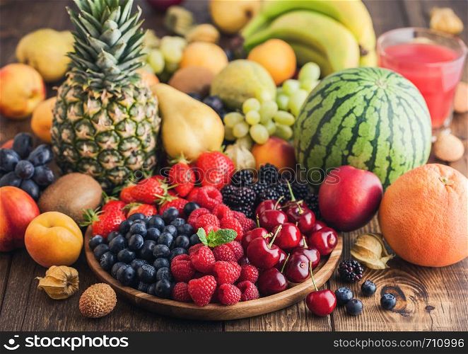 Fresh organic summer berries mix in wooden tray and exotic fruits on wooden background. Raspberries, strawberries, blueberries, blackberries and cherries. Watermelon, pear, pineapple, grapes. Top view