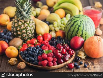 Fresh organic summer berries mix in wooden tray and exotic fruits on wooden background. Raspberries, strawberries, blueberries, blackberries and cherries. Watermelon, pear, pineapple, grapes. Top view