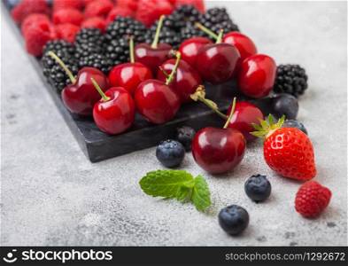 Fresh organic summer berries mix on black marble board on light kitchen table background. Raspberries, strawberries, blueberries, blackberries and cherries. Close up. Macro
