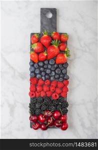 Fresh organic summer berries mix on black marble board on marble background. Raspberries, strawberries, blueberries, blackberries and cherries. Top view