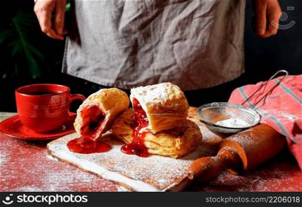 Fresh Puff staffed with plum or red currant jam on the table with a red cup and jar of jam on the black background.. Puff pastry dessert red jam viburnum cherry cup coffee black background