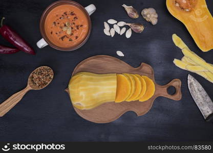 fresh pumpkin sliced into pieces on a wooden kitchen board and ingredients for cooking cream soup on a black background, top view