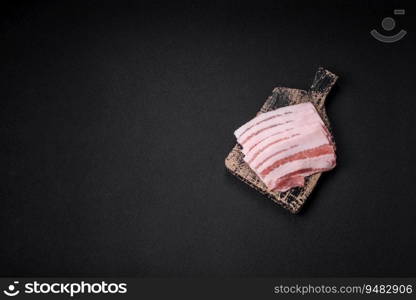 Fresh raw bacon cut into slices with salt, spices and herbs on a dark textured concrete background