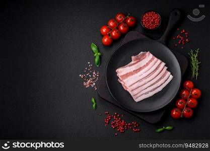 Fresh raw bacon cut into slices with salt, spices and herbs on a dark textured concrete background