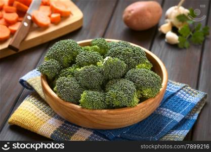 Fresh raw broccoli florets in wooden bowl with carrot slices on wooden board, potato, garlic and parsley in the back, photographed on dark wood with natural light (Selective Focus, Focus one third into the broccoli)