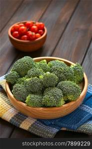 Fresh raw broccoli florets in wooden bowl with cherry tomatoes in the back, photographed on dark wood with natural light (Selective Focus, Focus one third into the broccoli)