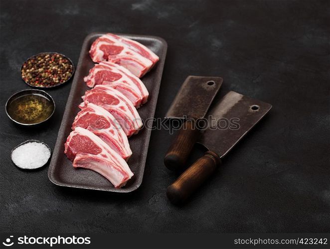 Fresh raw butchers lamb beef cutlets in plastic tray with vintage meat hatchets on black background.Salt, pepper and oil
