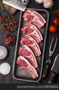 Fresh raw butchers lamb beef cutlets in plastic tray with vintage meat hatchet and fork with knife on black background.Salt, pepper, tomatoes and garlic.