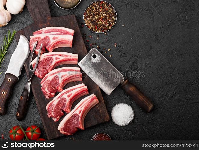 Fresh raw butchers lamb beef cutlets on stone board with vintage meat fork and knife and hatchet on black background.Salt, pepper and oil with tomatoes and garlic and barbecue sauce.