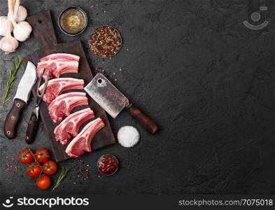 Fresh raw butchers lamb beef cutlets on stone board with vintage meat fork and knife and hatchet on black background.Salt, pepper and oil with tomatoes and garlic and barbecue sauce.
