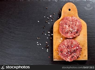 Fresh raw home-made minced beef steak burger with spices, on a cutting board on black slate table, copy space, top view. raw home-made beef burger cutlet