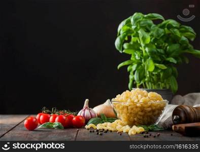 Fresh raw maccherono elbows pasta in glass bowl with basil plant, oil and tomatoes with garlic and pepper on wooden table background. Space for text