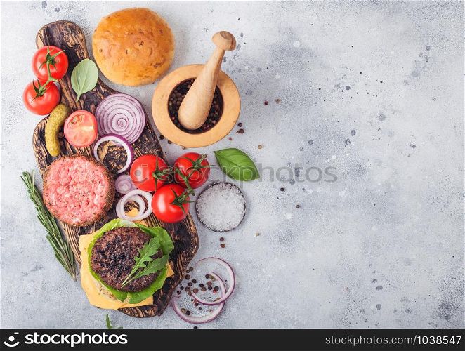 Fresh raw minced and grilled pepper beef burgers on vintage chopping board with buns onion and tomatoes on wooden background.Mortar with pestle with pickles and basil. Space for text.