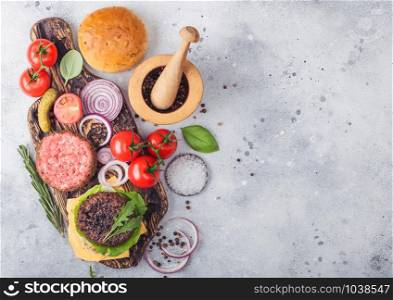 Fresh raw minced and grilled pepper beef burgers on vintage chopping board with buns onion and tomatoes on wooden background.Mortar with pestle with pickles and basil. Space for text.