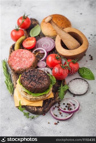 Fresh raw minced and grilled pepper beef burgers on vintage chopping board with buns onion and tomatoes on wooden background.Mortar with pestle with pickles and basil. Top view