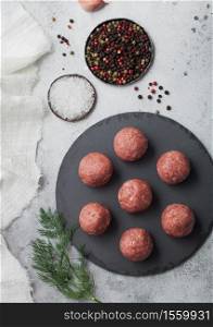 Fresh raw minced beef meatballs on round stone board with pepper, salt and garlic on light table background with dill and towel.